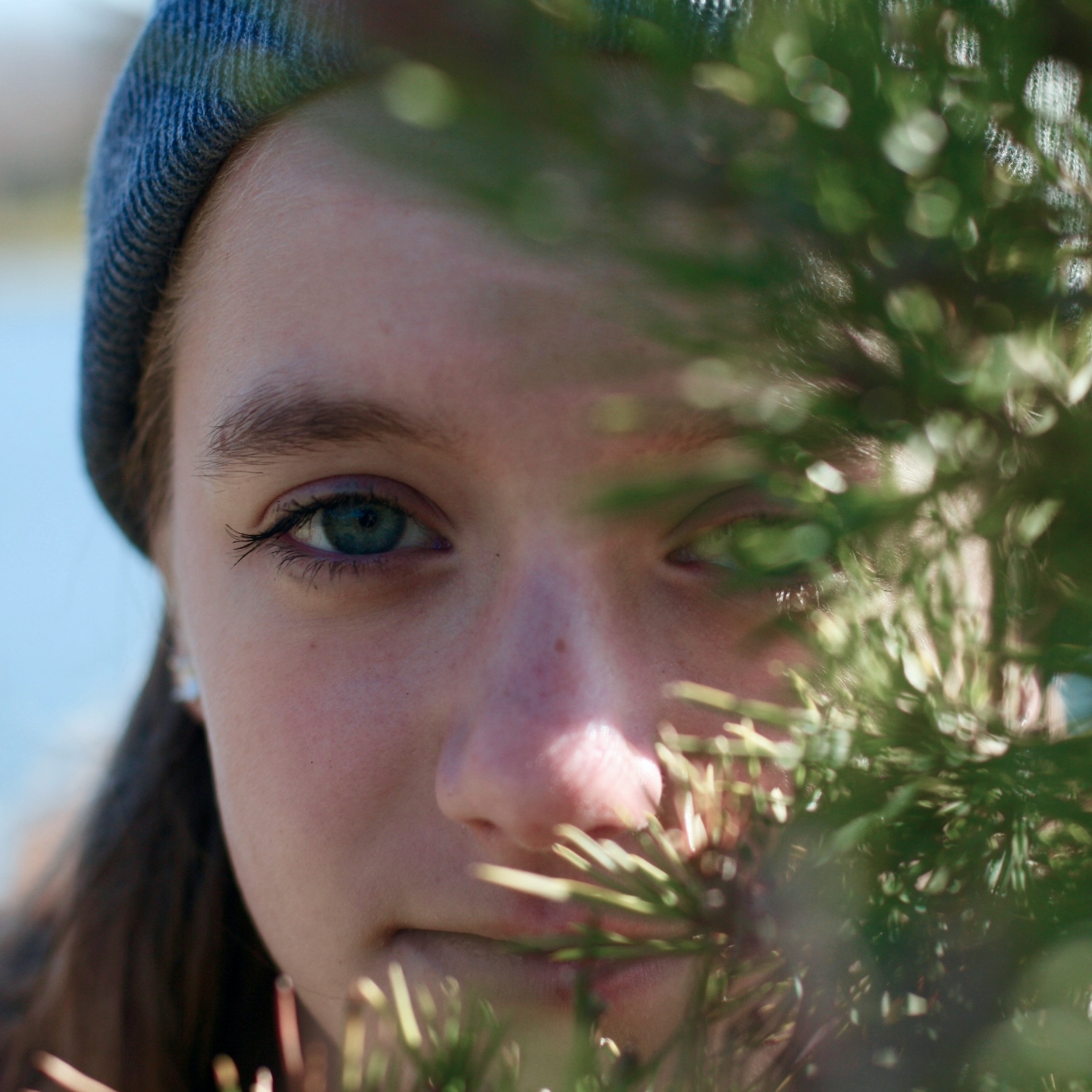 Close-up of a person partially obscured by pine branches, wearing a blue knit beanie
