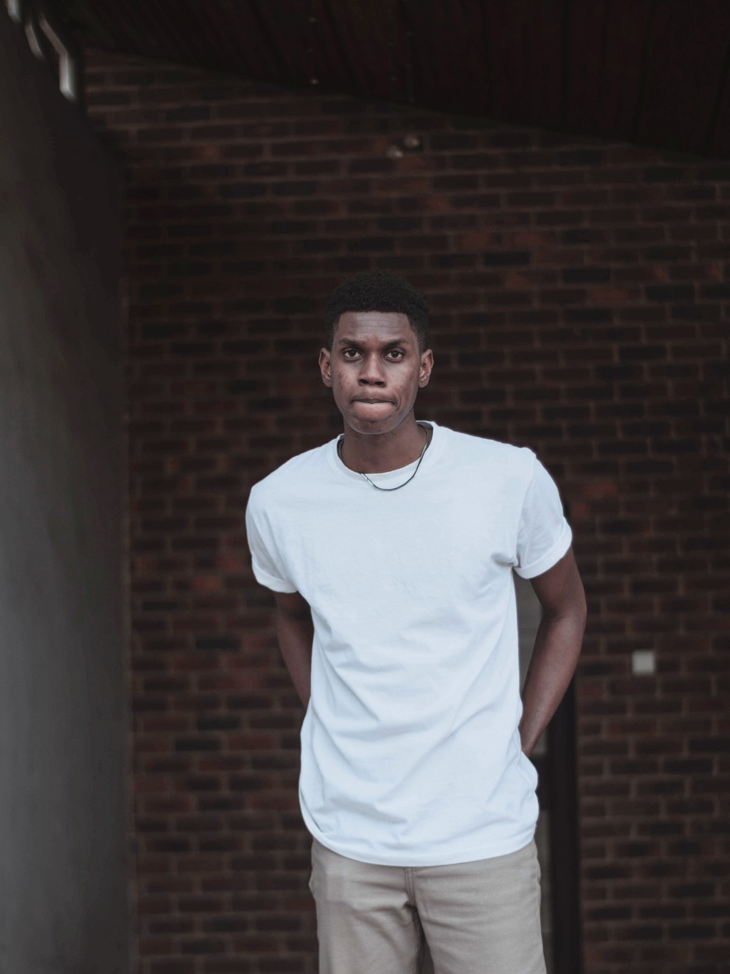 Young man in a white t-shirt standing in a brick archway, hands in pockets, facing the camera