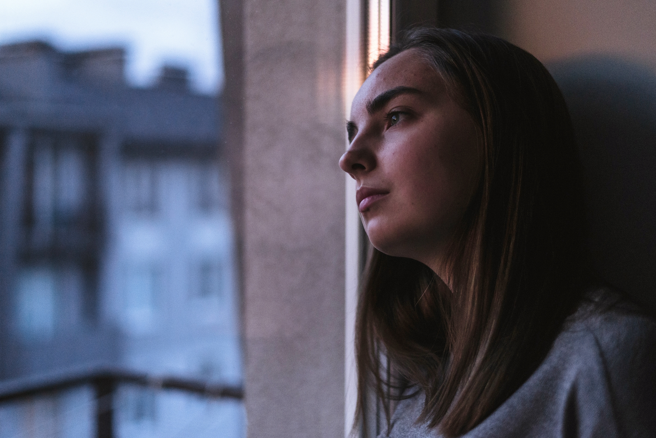 Young woman sitting indoors gazing out a window, with natural light casting a soft glow on her thoughtful expression