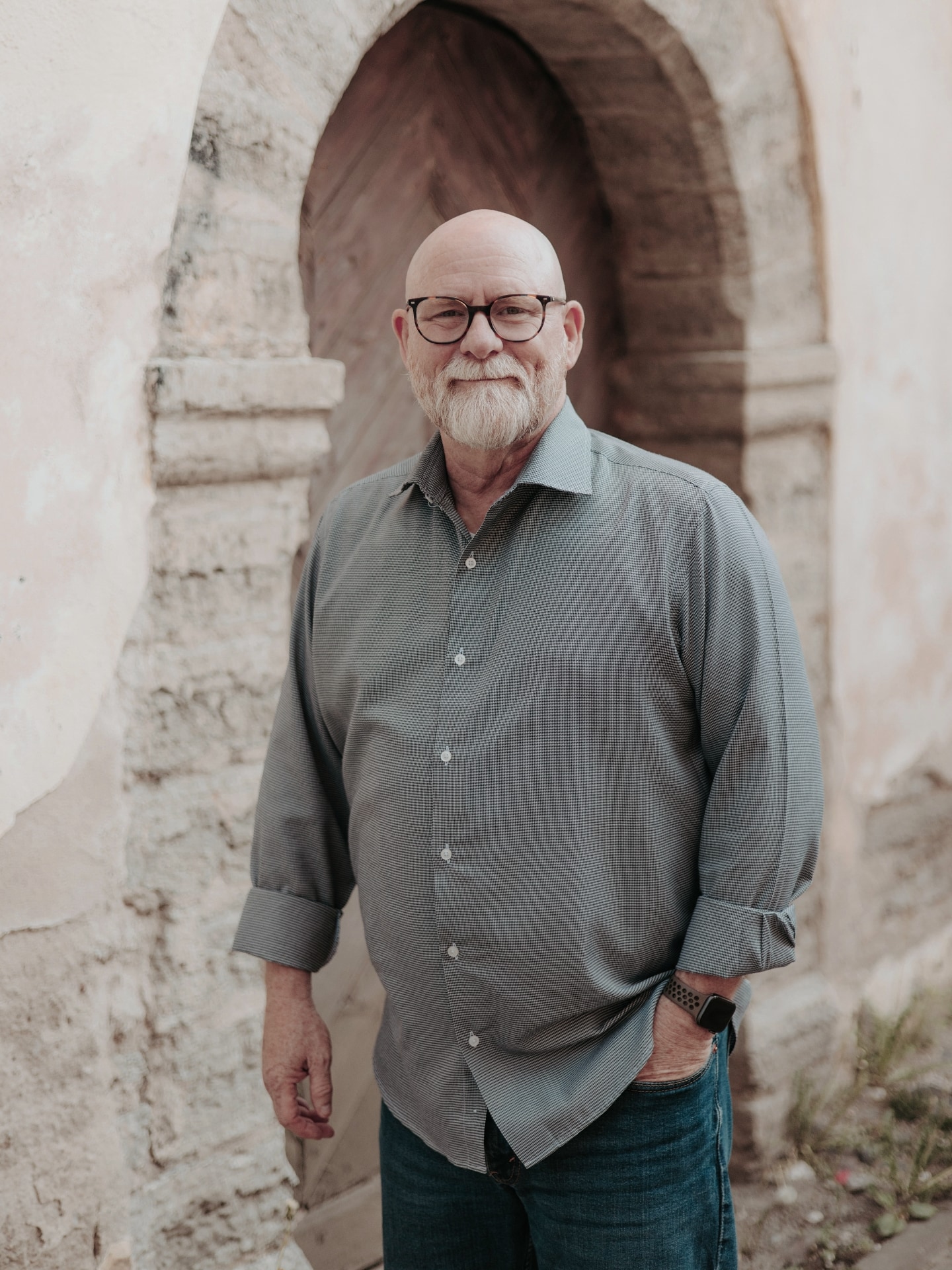 Portrait of a bald man with glasses, a beard, and a warm smile, standing casually in front of a textured archway in an outdoor setting, wearing a button-up shirt and jeans