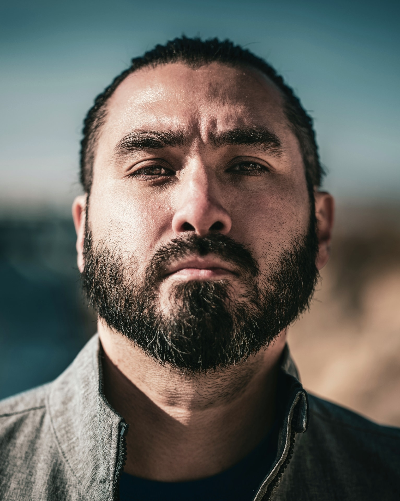 Close-up portrait of a bearded man wearing a dark jacket against a softly blurred natural background
