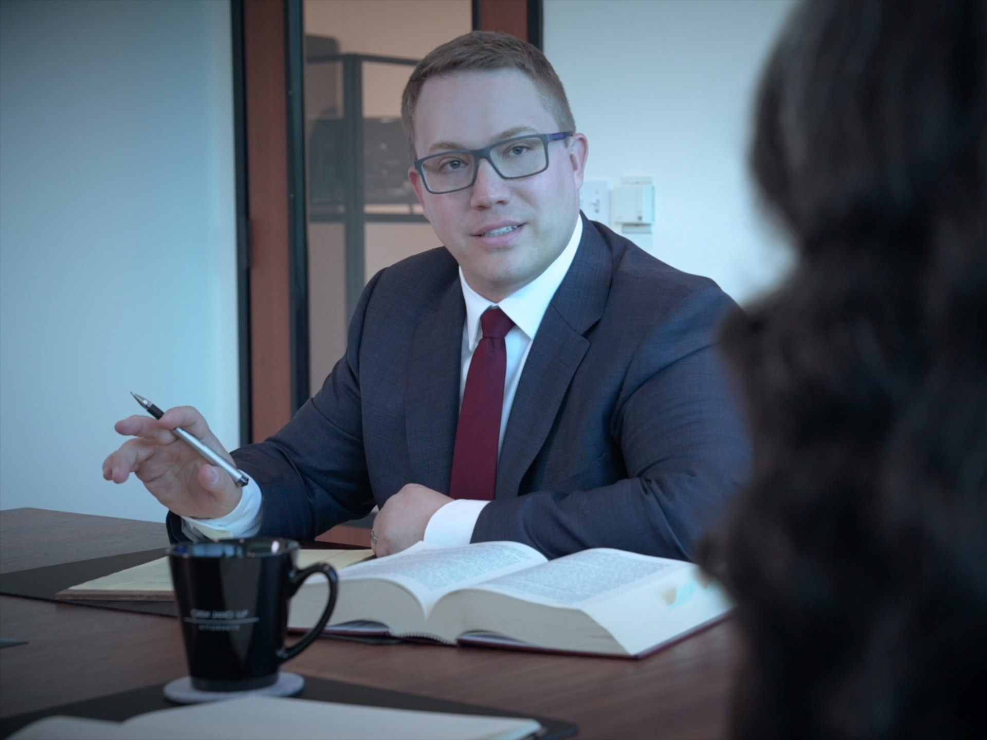 Attorney in a navy suit and burgundy tie seated at a conference table, holding a pen over an open law book with a Crew Janci mug and notebook nearby, listening to a colleague whose back is to the camera
