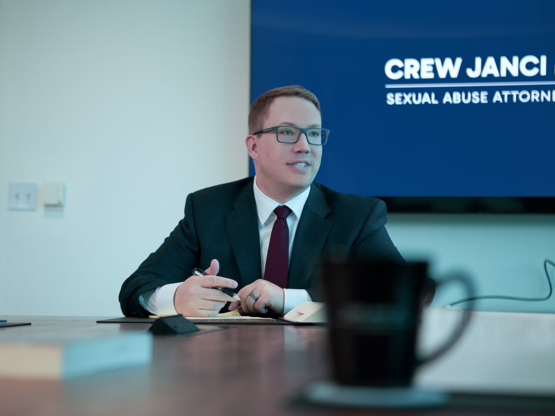 Attorney Peter Janci in a suit and tie speaking at a conference table, with a screen in the background displaying the words ‘Crew Janci Sexual Abuse Attorneys'