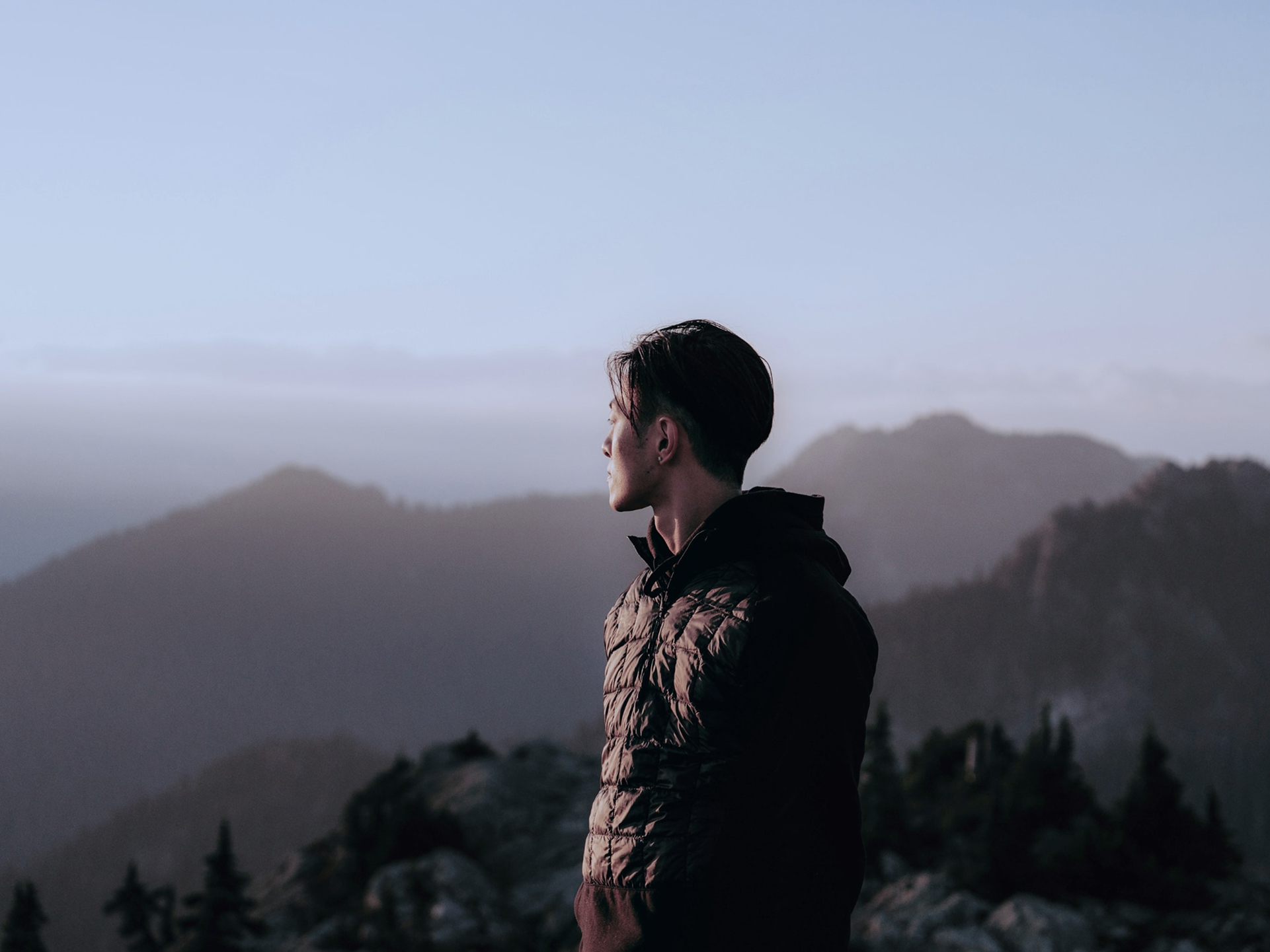 Man standing on a mountain overlook at dusk, gazing toward the horizon with layers of misty peaks in the distance